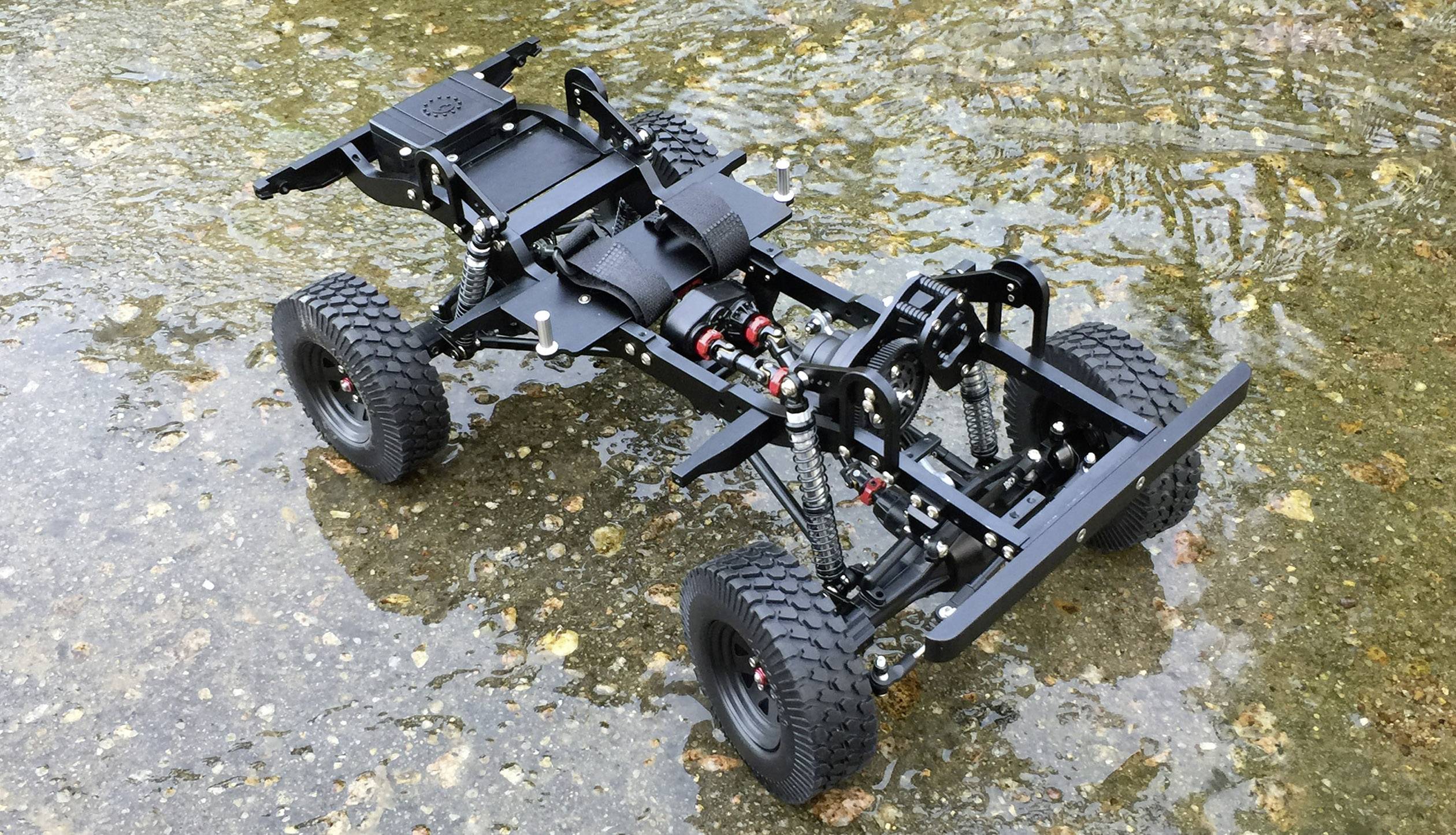 A model car chassis with 4 large tyres and visible suspension stands on wet, gravelly ground.