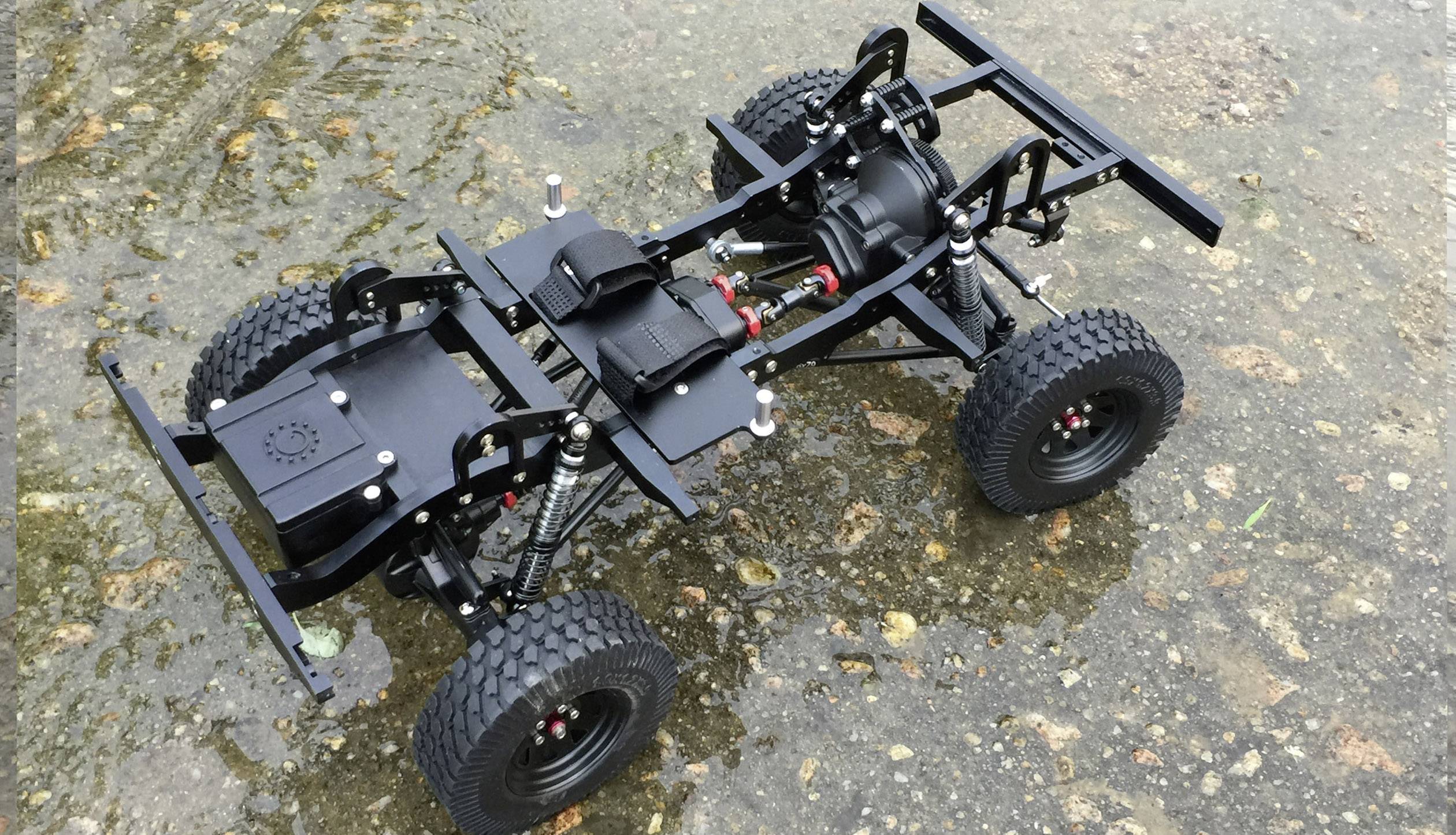 A remote-controlled car chassis is resting on a wet surface. It reveals the suspension, tyres, and inner structure of the vehicle.