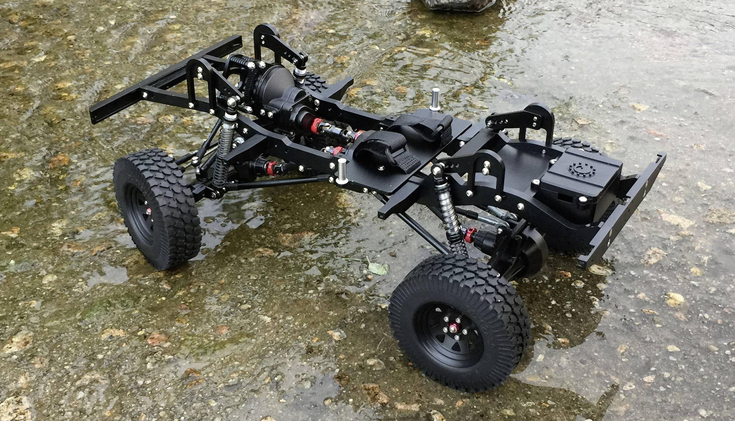 A remote-controlled vehicle chassis with rugged off-road tyres and visible suspension components is standing on a wet, rocky surface.