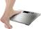 A person places one foot on a digital body scales with a stainless steel surface. The scales display two zeros on the screen.