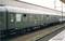 A green passenger train carriage is standing on a railway track. It has several windows and the inscription '2nd Class'. The background shows a railway station building.