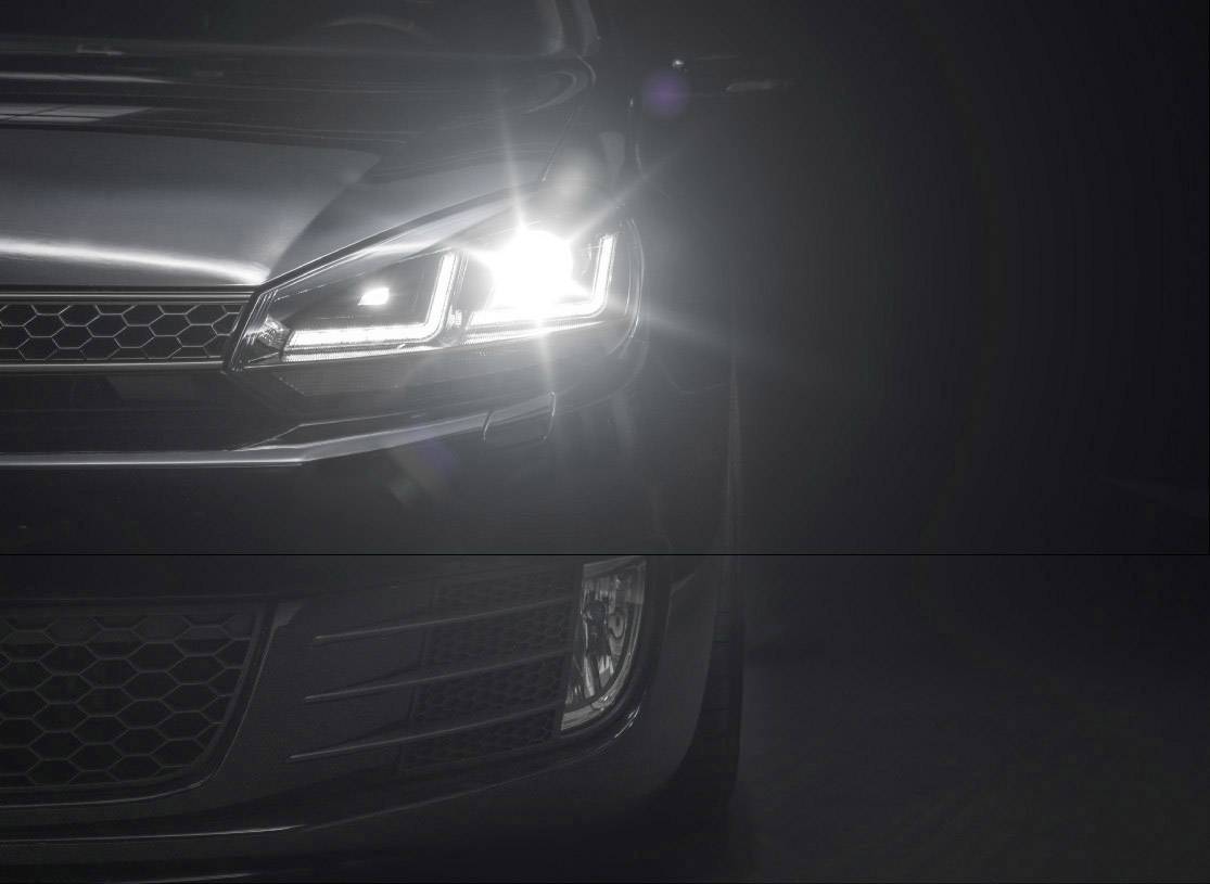 A car with its headlight switched on in the dark. The right headlight is brightly illuminated and creates a beam of light.