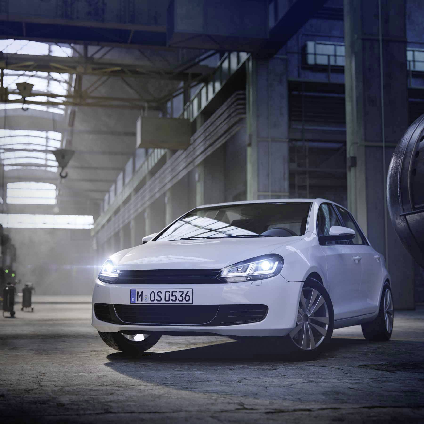 A white car is parked in a spacious, well-lit industrial hall, where light streams through the glass roof, casting shadows.