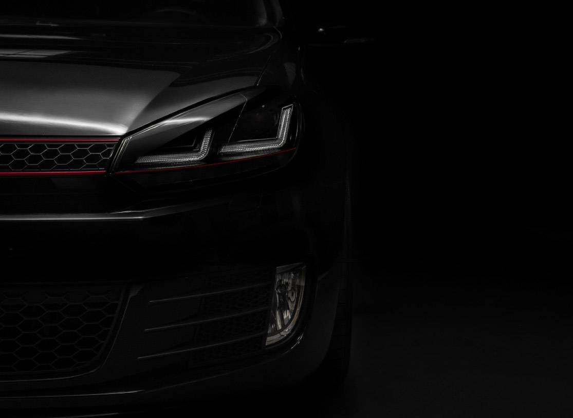 Half profile of a black car in dim light, showing the front headlight and part of the radiator grille.