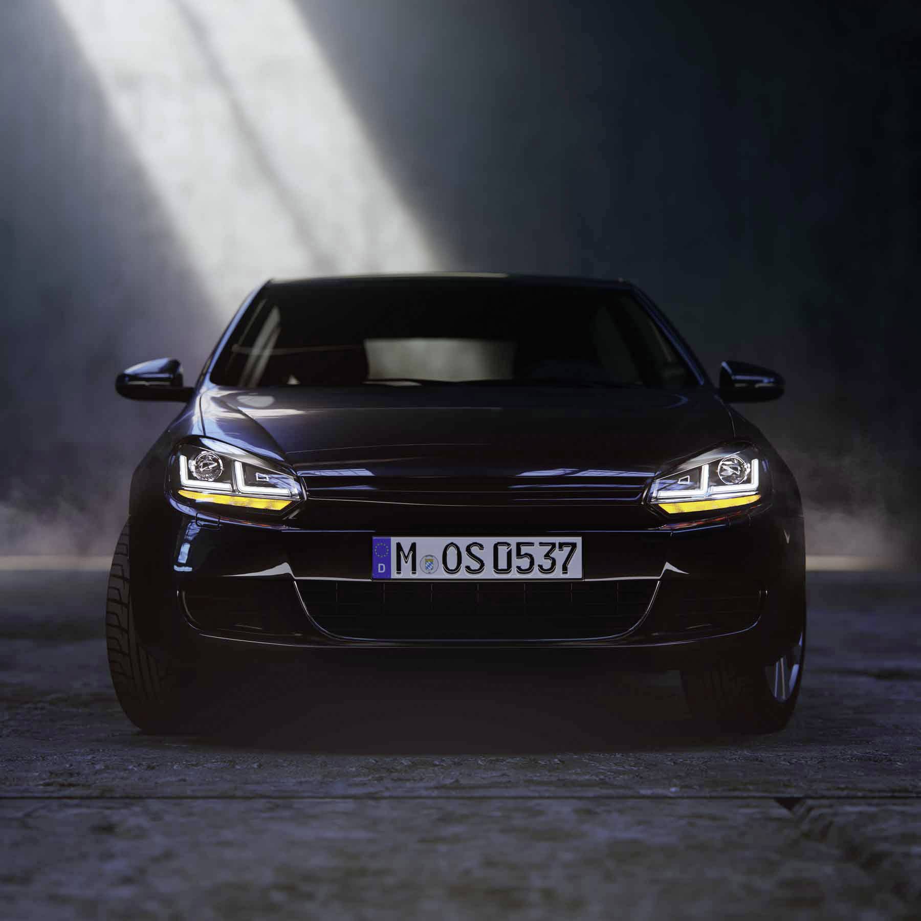 Black car with headlights on stands on a concrete surface, viewed from the front, with a beam of light from above.