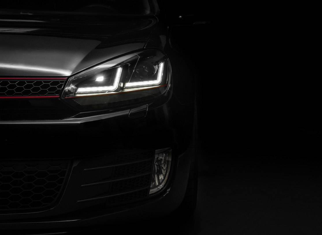 Front view of a car with its headlight switched on and a red stripe on the radiator grille, against a dark background.