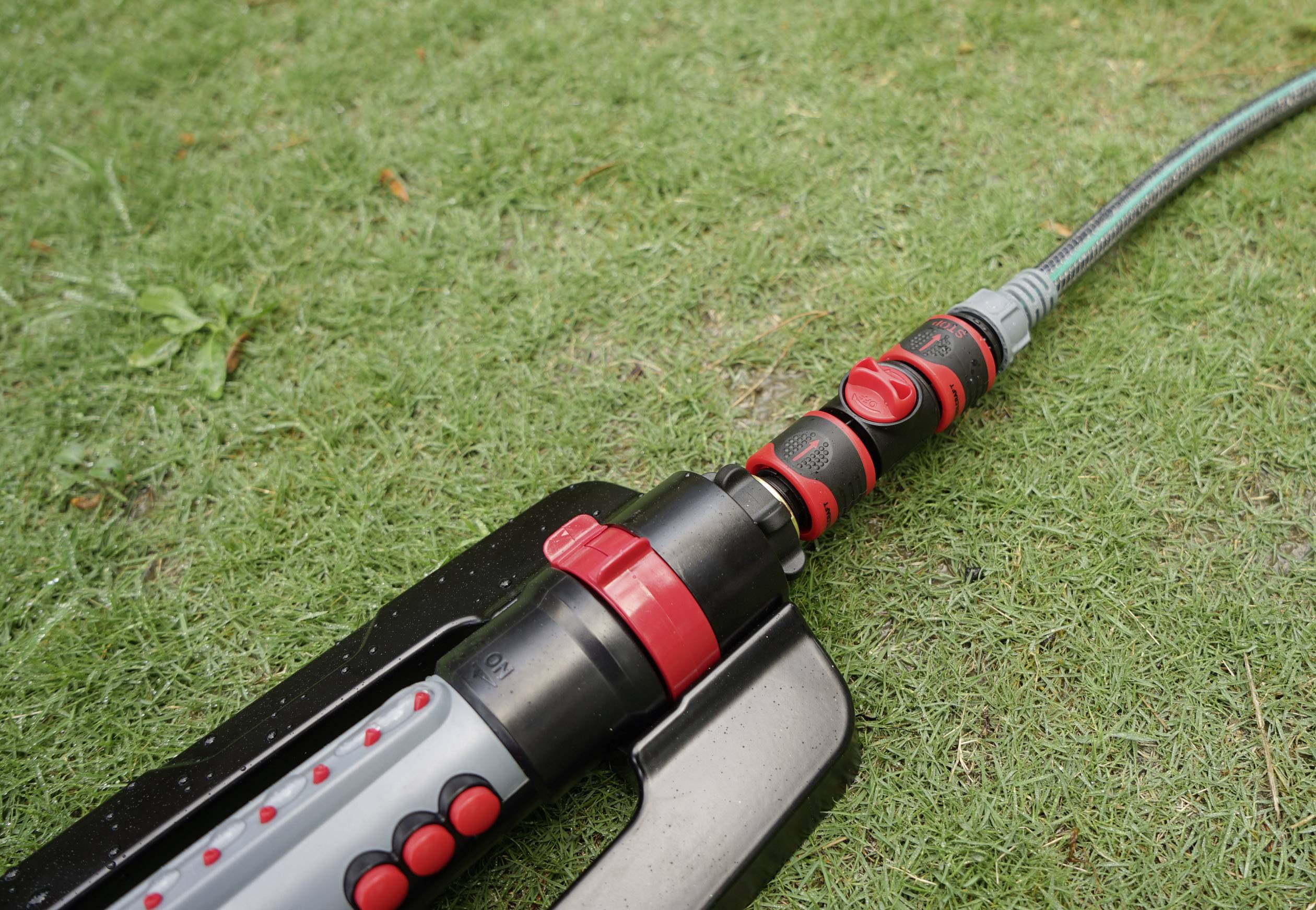 Red and black lawn sprinkler on green grass, connected to a garden hose.<br><br>Note: This is an exact translation maintaining the original description. In British English, the terminology remains the same, as this is a straightforward technical description.