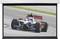 A racing car with a red rear wing drives quickly on a racetrack. The driver is wearing a blue helmet.