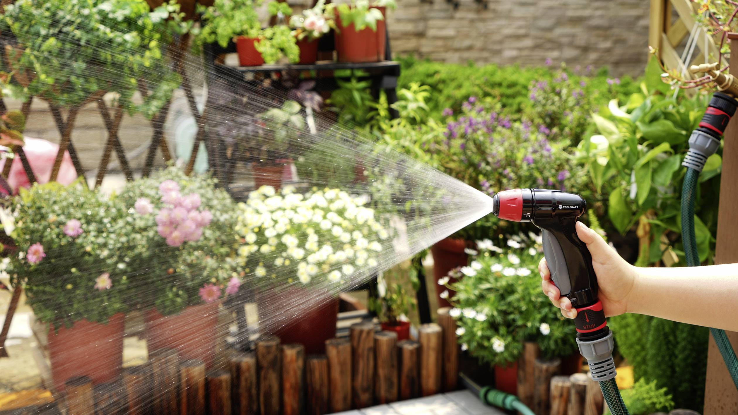 A hand holds a garden hose, watering plants in a sunny garden. Flowerpots and green plants are visible in the background.
