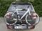 A white bicycle is attached to a bike rack at the rear of a black car, which is parked on a paved car park.