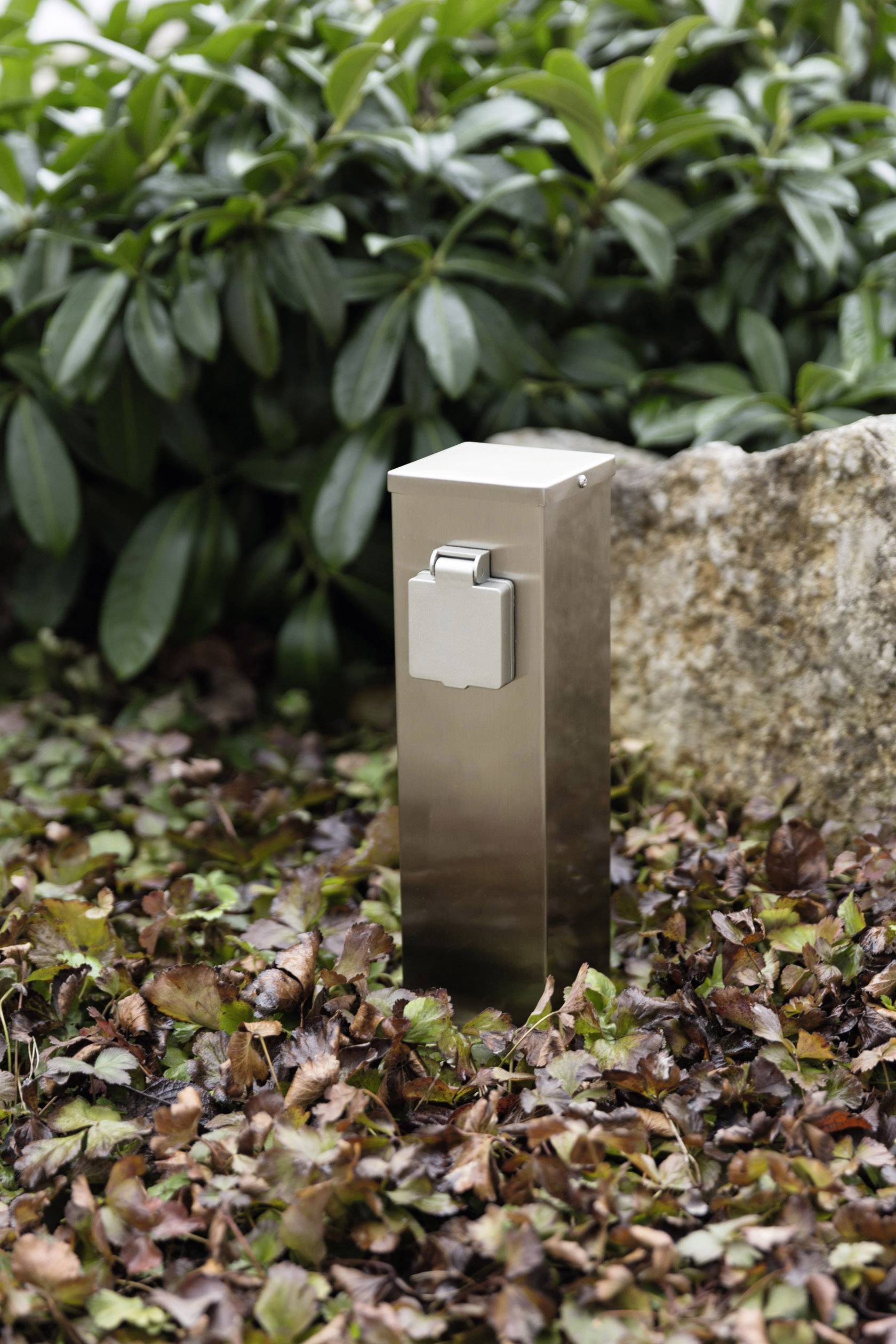 Metal outdoor socket, surrounded by leaves and shrubs, suitable for external areas such as gardens or patios.