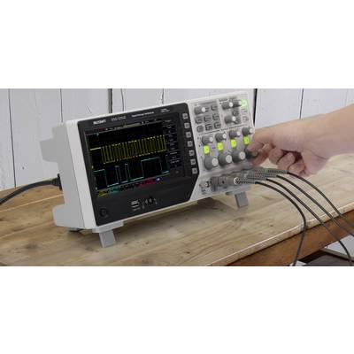 A person is operating an oscilloscope on a wooden table, displaying electrical signals with waveforms on a screen.