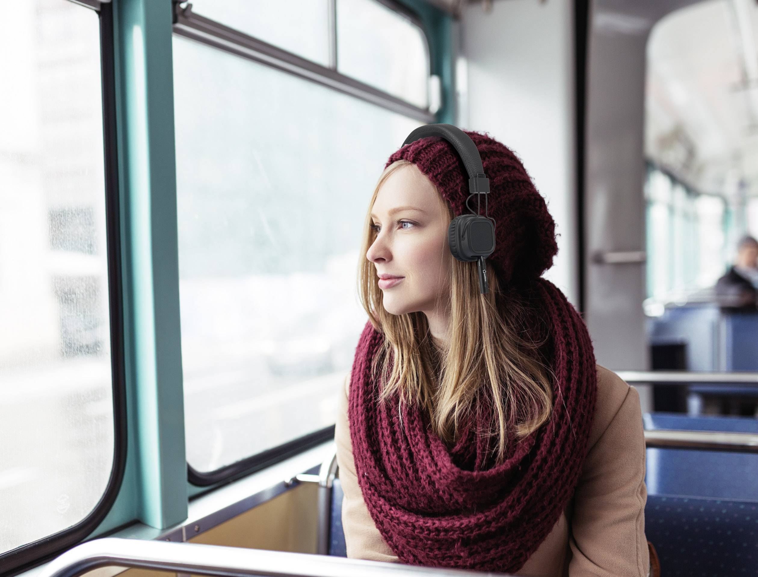 A woman wearing a hat and headphones looks out of a bus window. She is wearing a red scarf and sits relaxed.