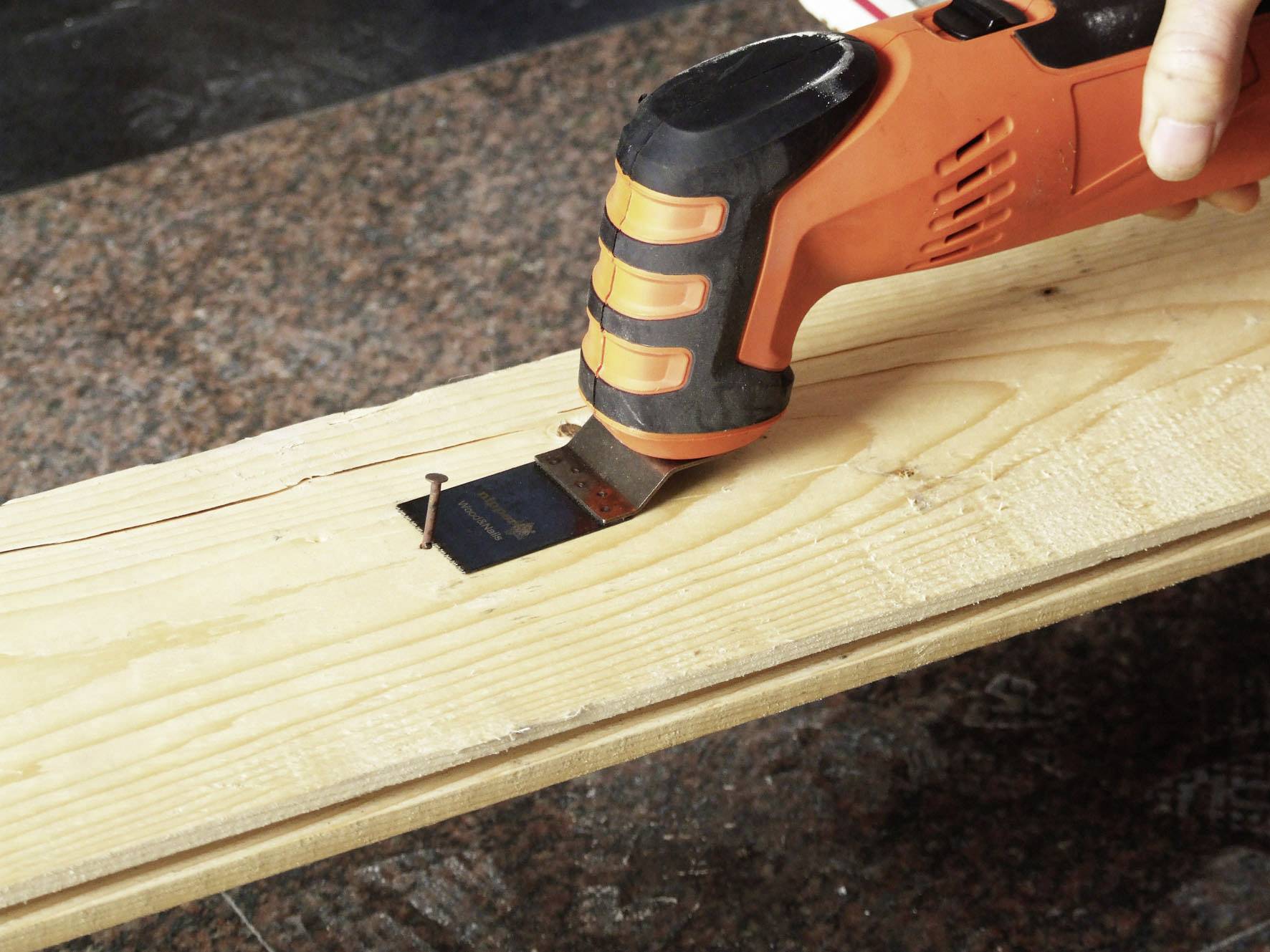 A hand is holding a multi-functional tool with an orange handle, which is working on a cut-out section in a wooden beam. A nail is embedded in the wood.