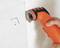 A hand is holding an orange electric tool and cutting a small hole in a white wall or tile.