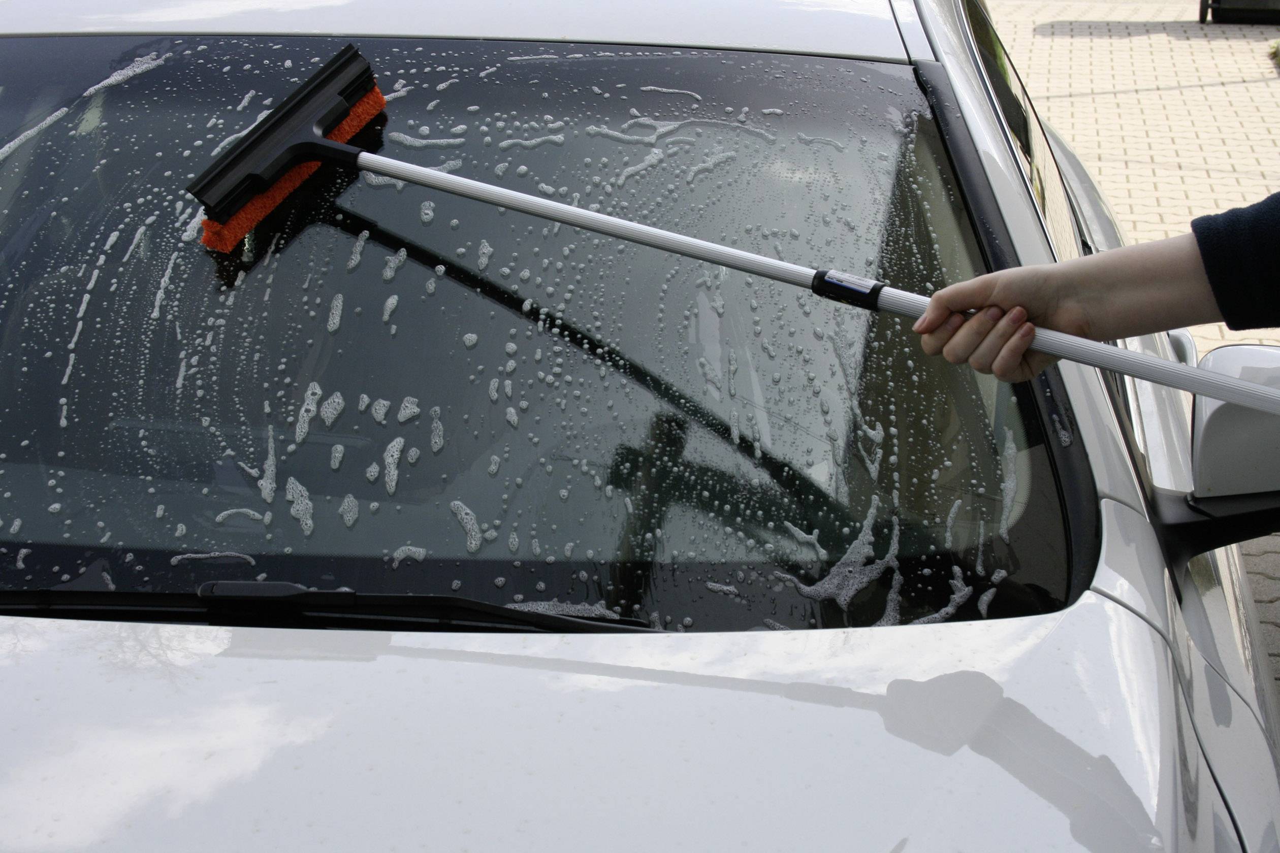 A person is cleaning the windscreen of a car with a long window wiper, with water and soap clearly visible. The car is parked in a car park.