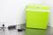 Small green cool box on a metal surface next to a power socket. A black power adapter is connected, with the cable running to the socket.