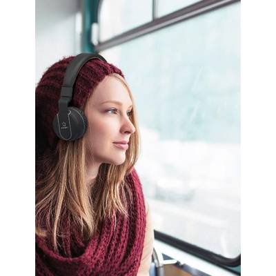 A woman wearing headphones sits on a bus and looks out of the window. She is wearing a red scarf and a hat. Exterior view blurred.