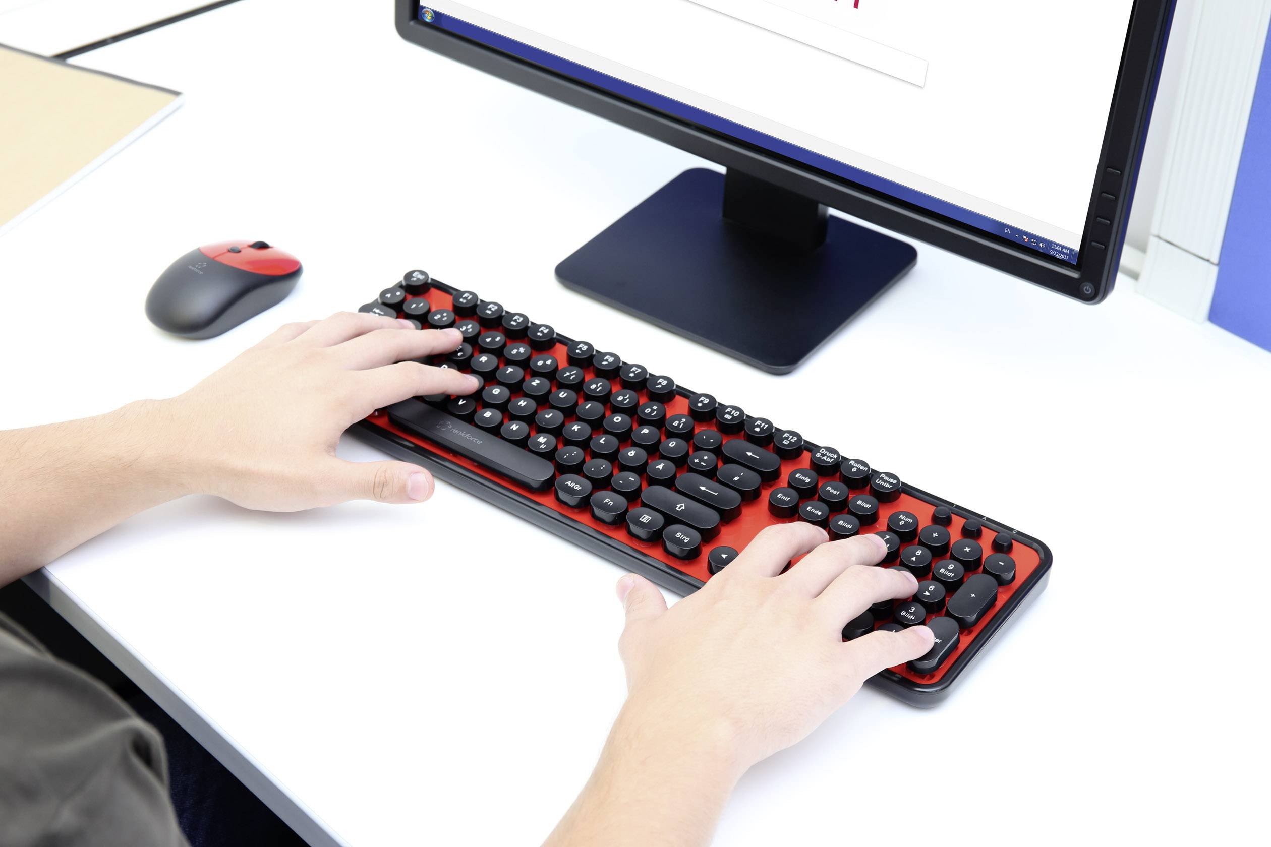 A person is typing on a red and black keyboard while looking at a monitor. The scene depicts working on a computer.