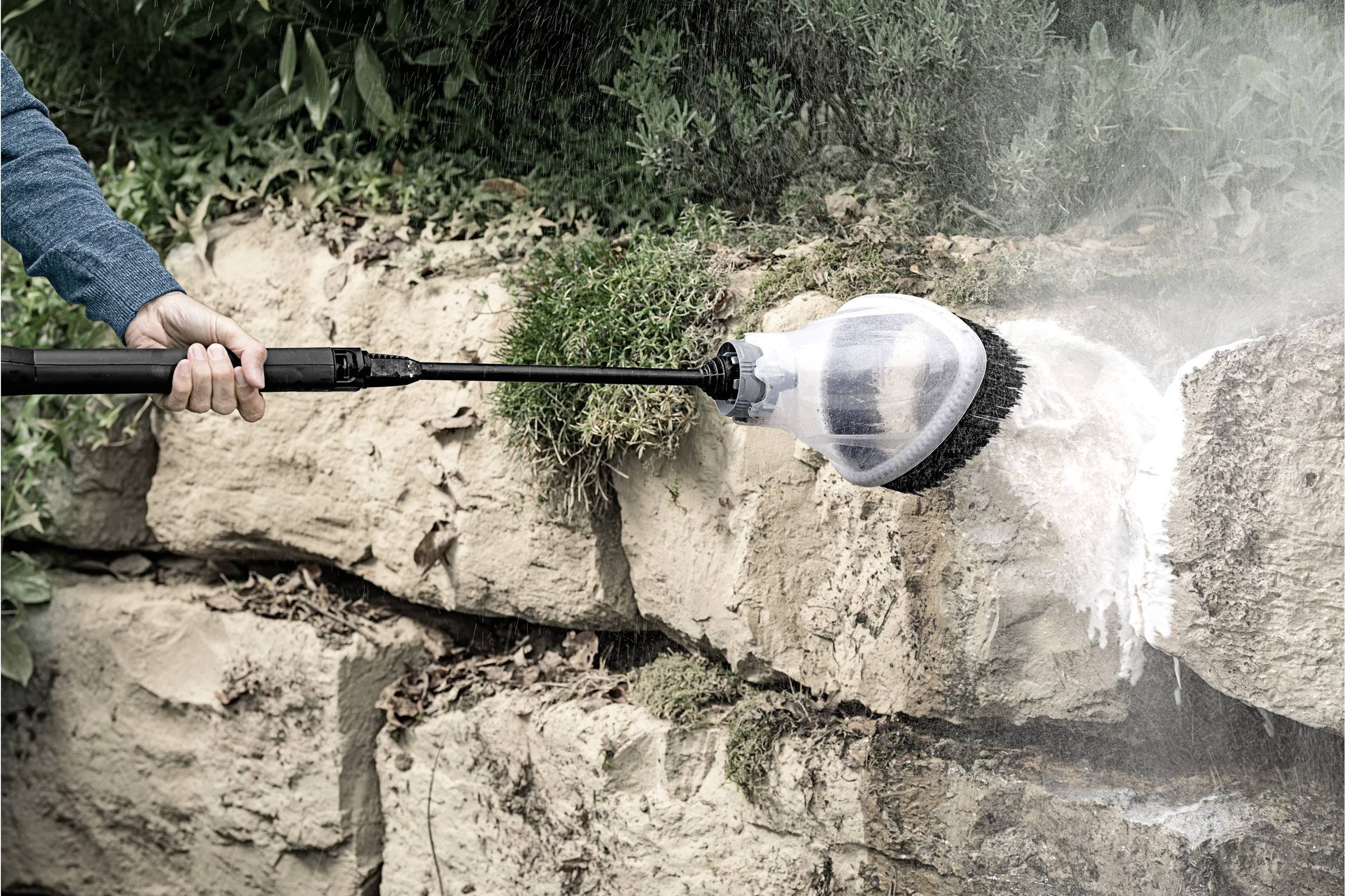 A pressure washer is cleaning rocks. A rotary brush removes dirt from a stone surface. Plants are visible in the background.