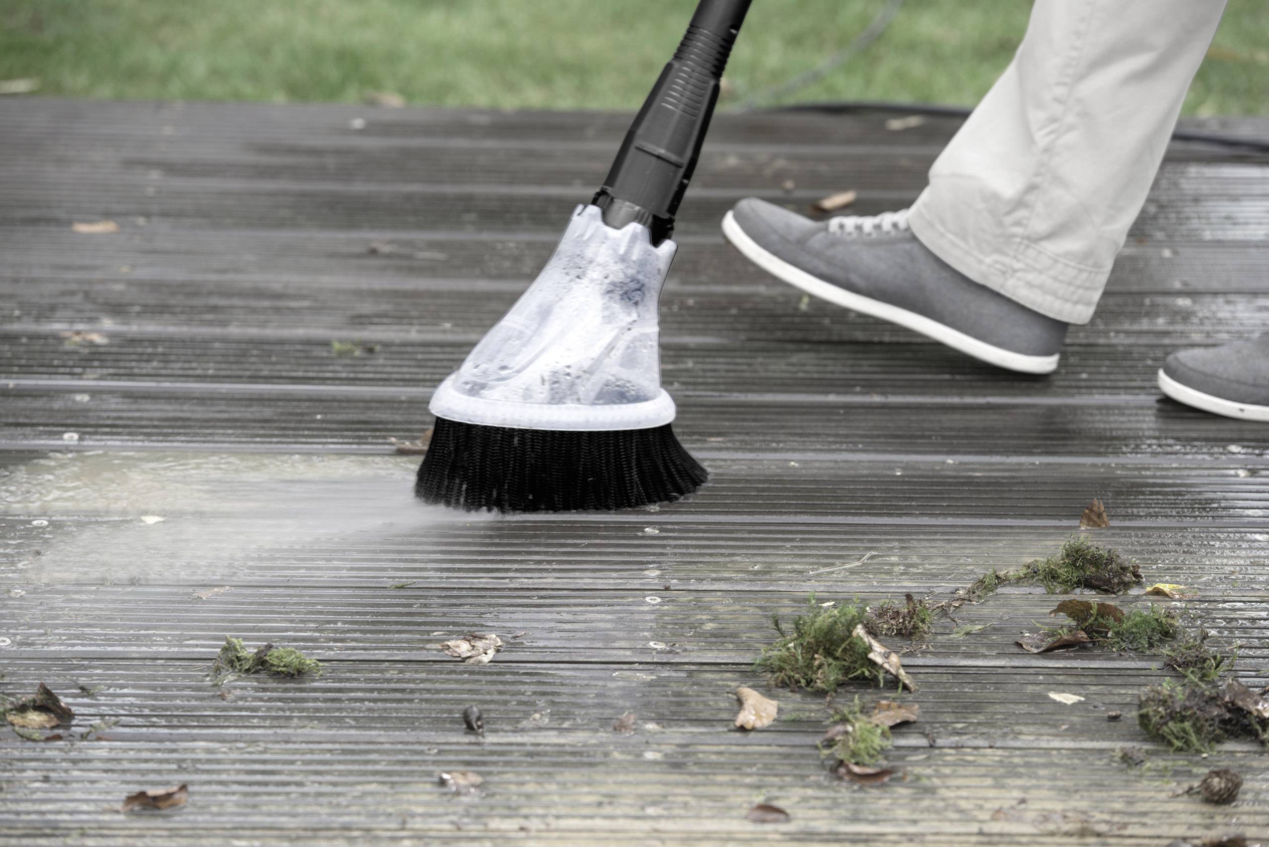 A person wearing grey shoes is cleaning a wooden terrace with a pressure washer. Leaves and moss are being removed.