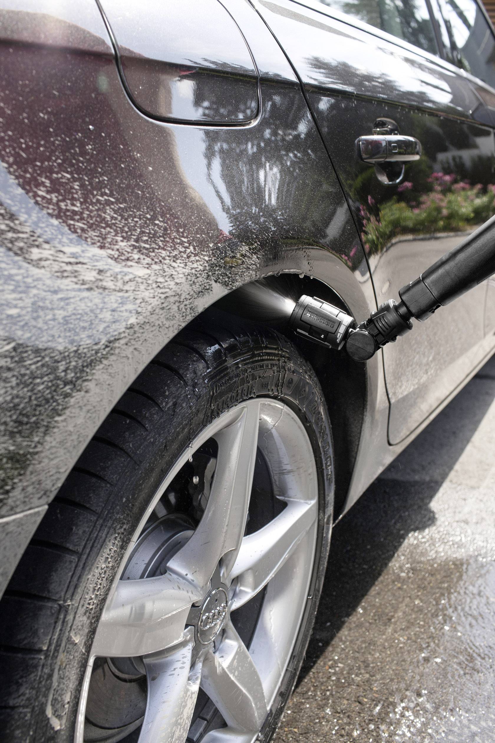 A car is being cleaned with a high-pressure washer at the wheel. Water droplets and foam are visible on the tyre and bodywork.