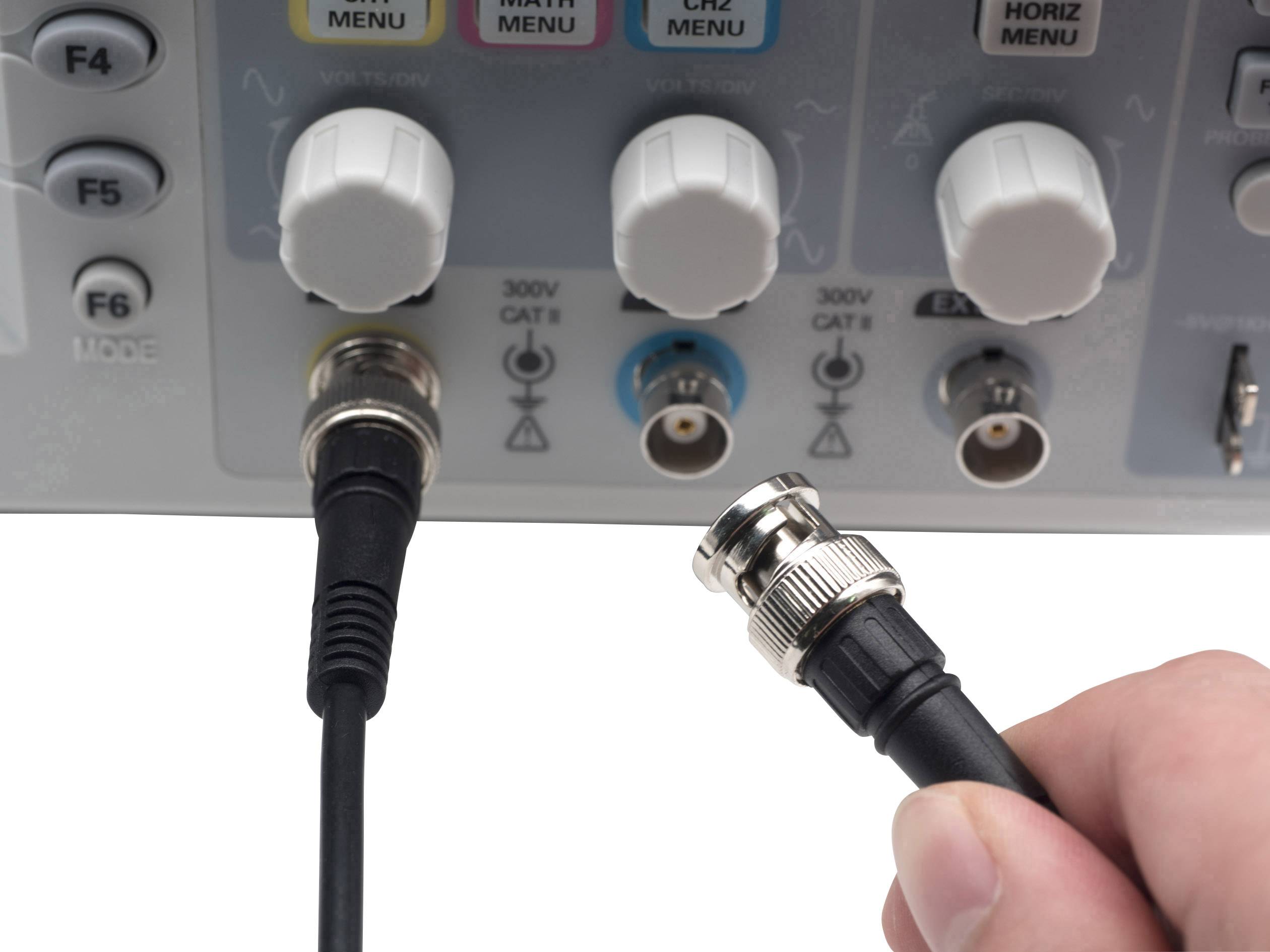 A hand holds a cable plug being connected to a measuring device with multiple rotary knobs. Symbols and connectors are visible.