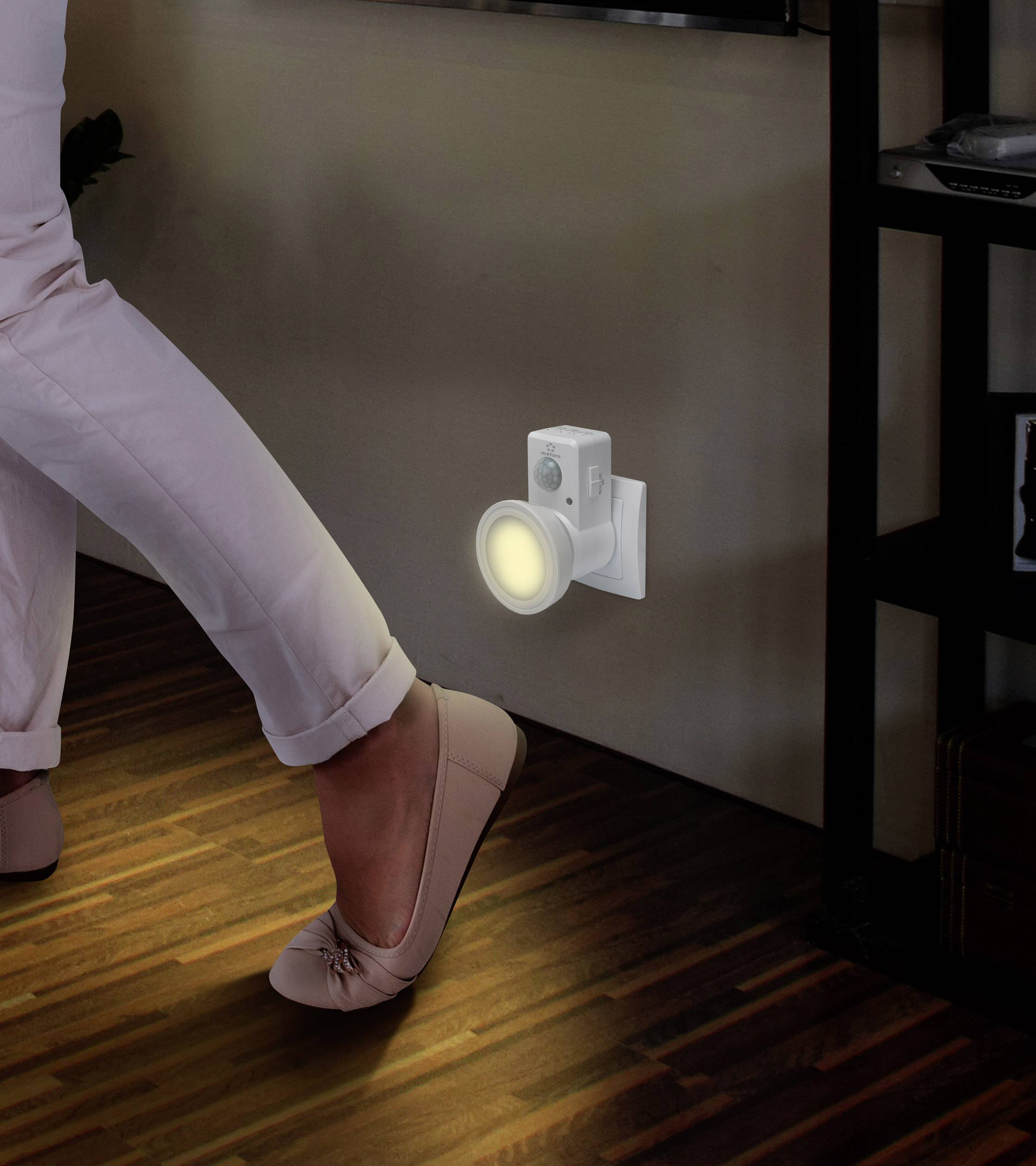 A person walks in a dark room; a night light with a motion sensor illuminates the floor and lights the way.