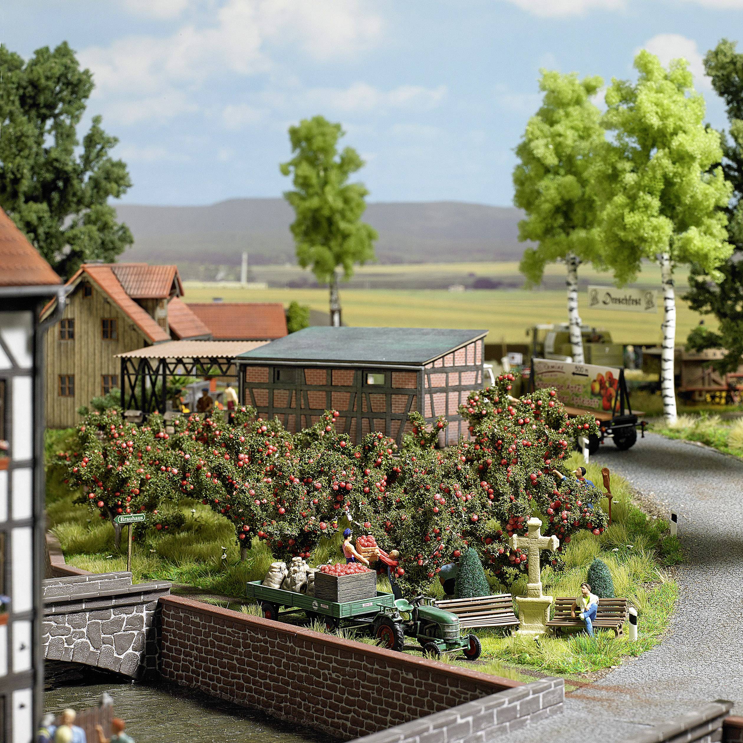 Model railway landscape with half-timbered houses, apple trees, and people. A bridge crosses a river. Figures are busy.