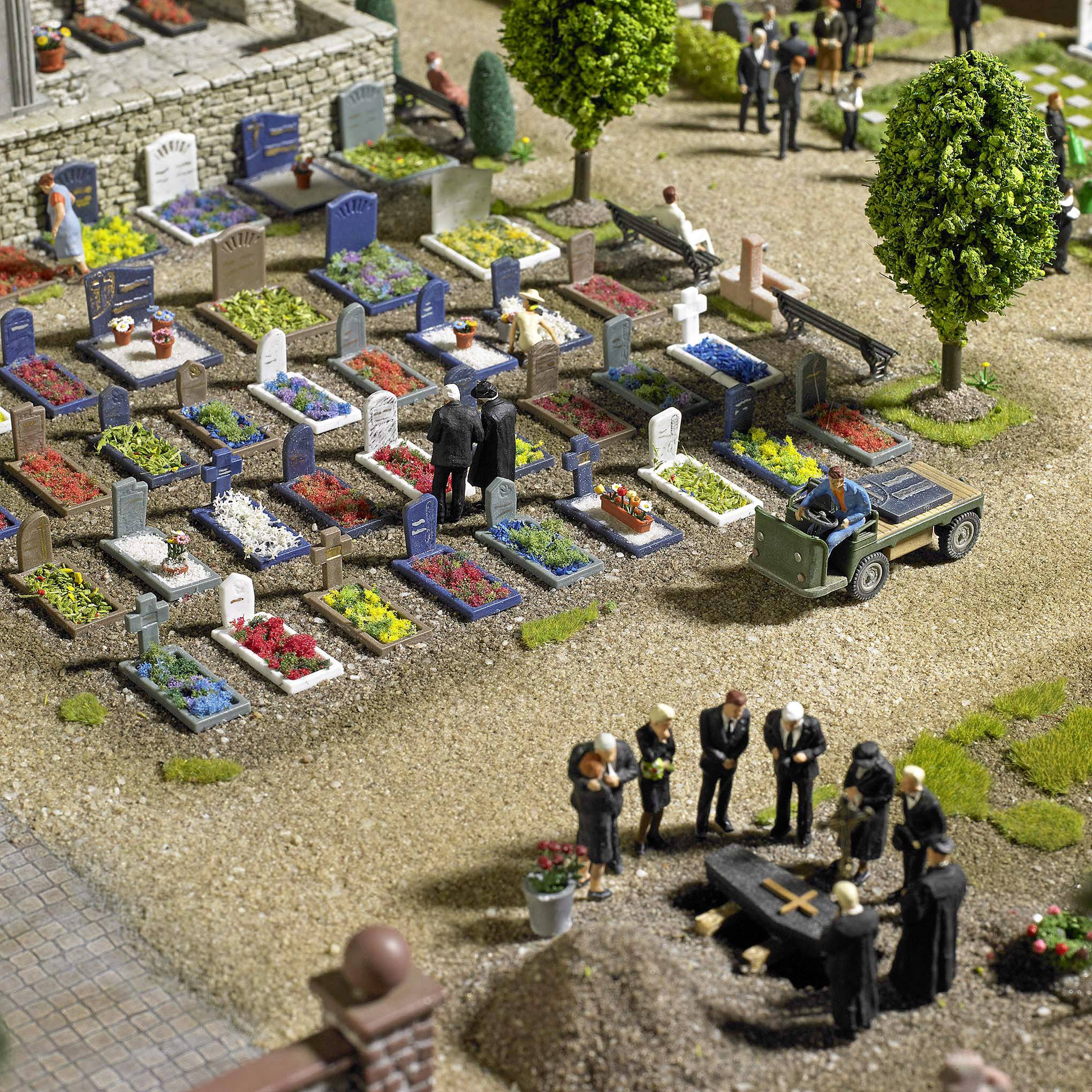 Miniature cemetery scene with figures at a funeral, several graves, a green hearse. Trees and flowers line the path.