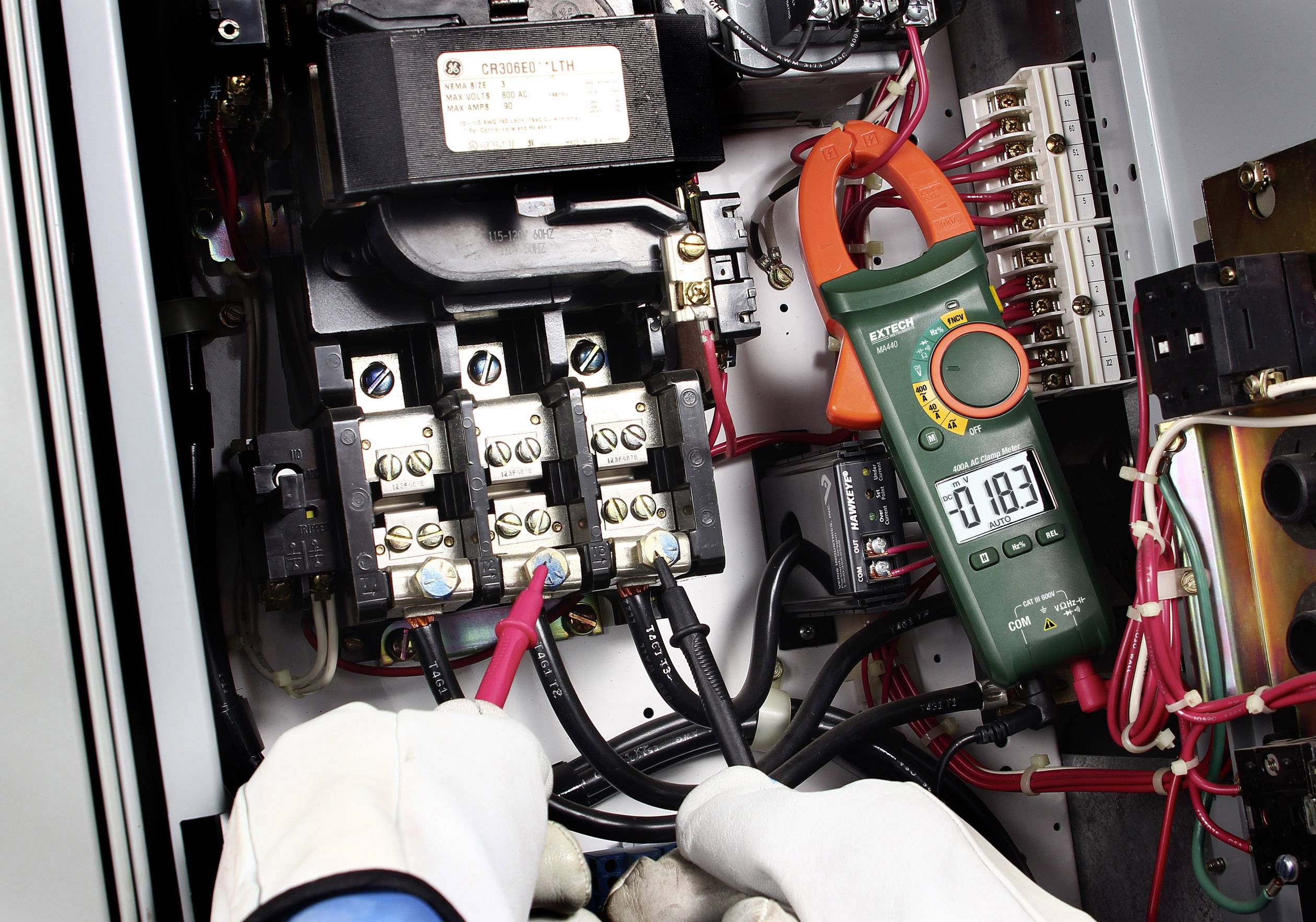 An electrician is measuring the electric current with a multimeter in a control panel. The display shows a value of 18.3.