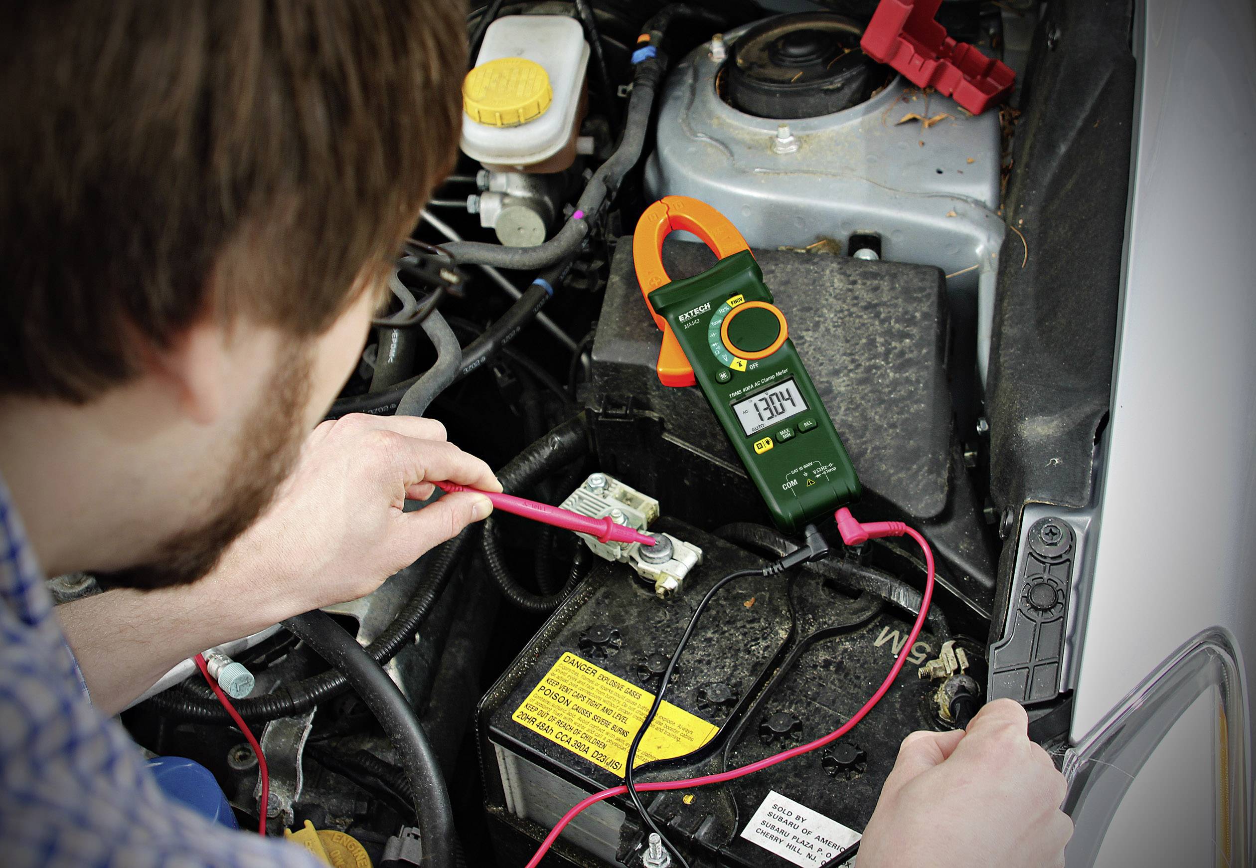 A man is measuring the voltage of a car battery with a multimetre. The display shows 13.08 volts, with red cables connected to the terminals.