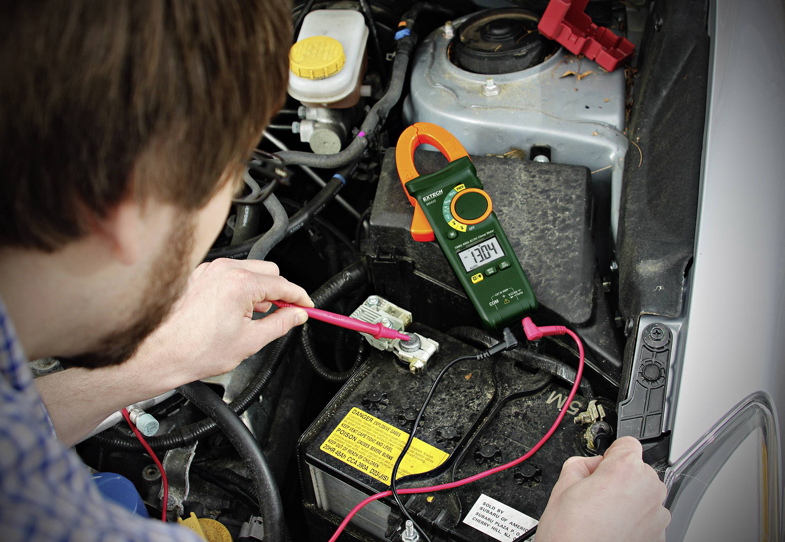 A man is checking the voltage of a car battery in the engine compartment using a multimeter. The device is showing 12.84 volts.