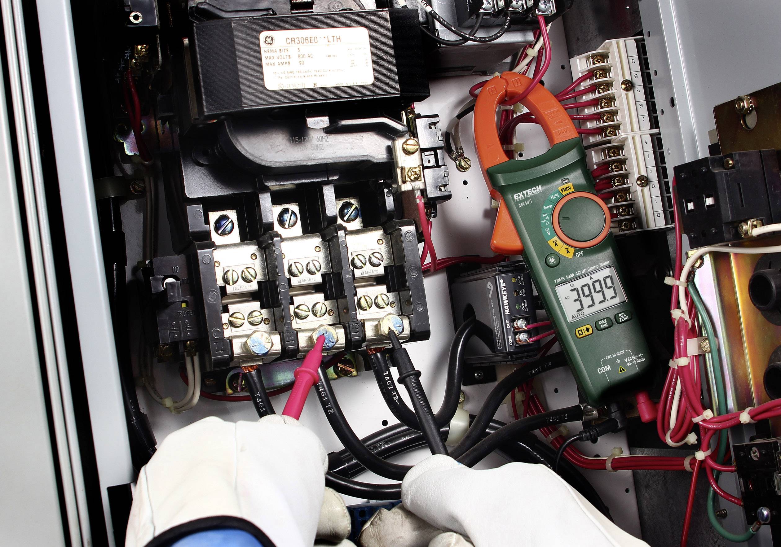'Hands wearing protective gloves measuring electrical voltage using a multimeter in an electrical cabinet. Display shows -3999.'