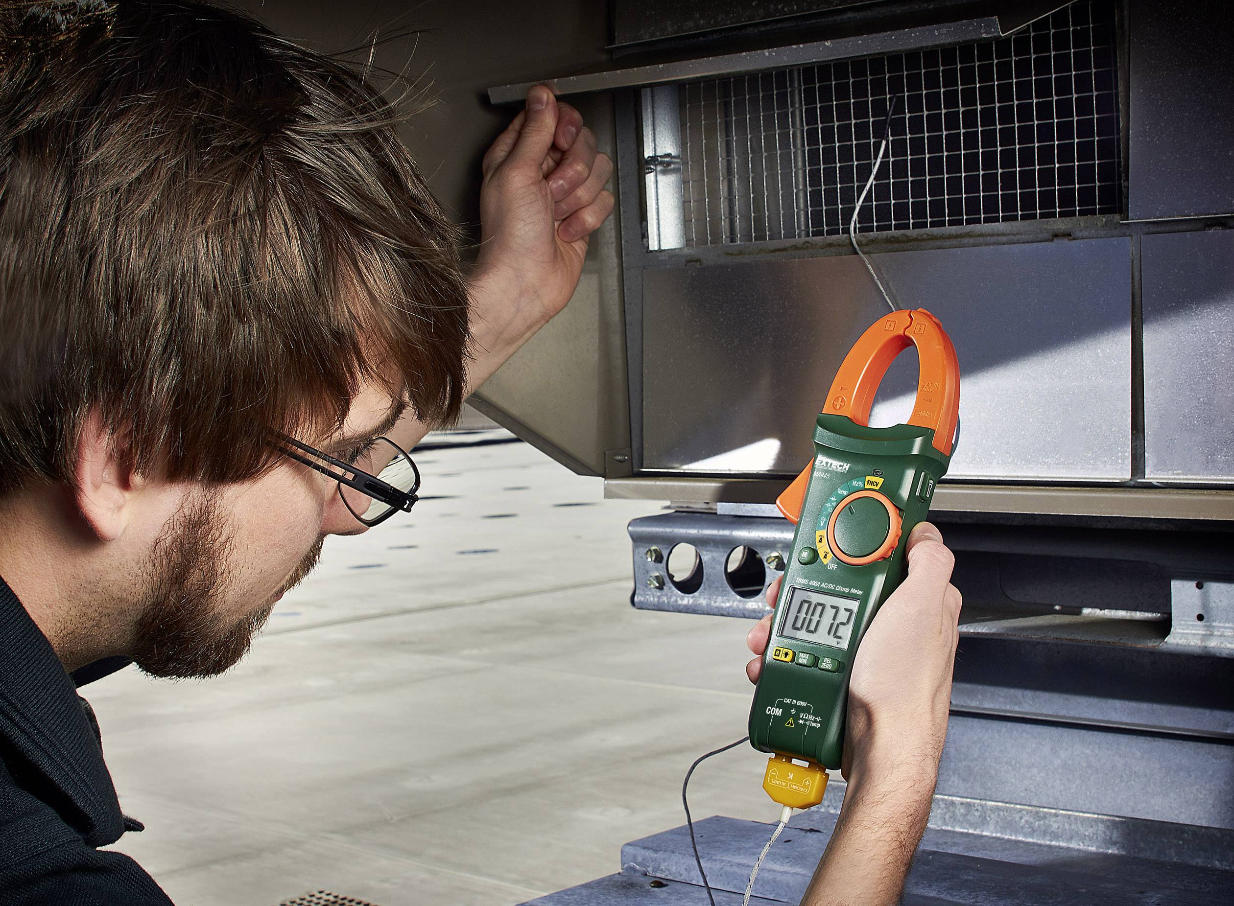 A man is measuring the voltage on an electrical device using a multimeter. He is wearing glasses.