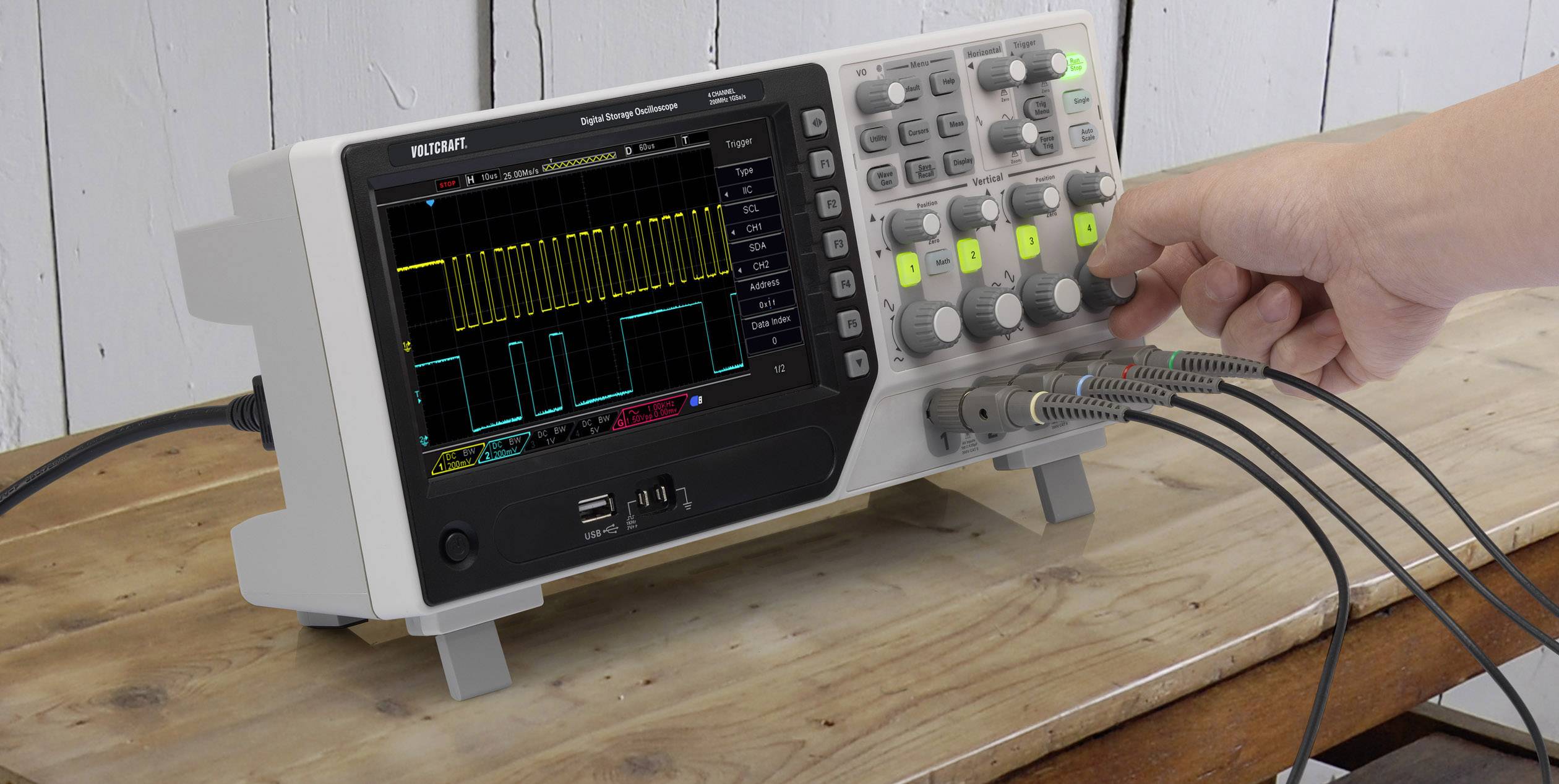 Digital storage oscilloscope on a wooden table showing rectangular waves. A hand operates the controls to adjust the display.
