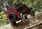 A robust, off-road model car drives over uneven, wooded terrain. Large tyres and a distinctive design.