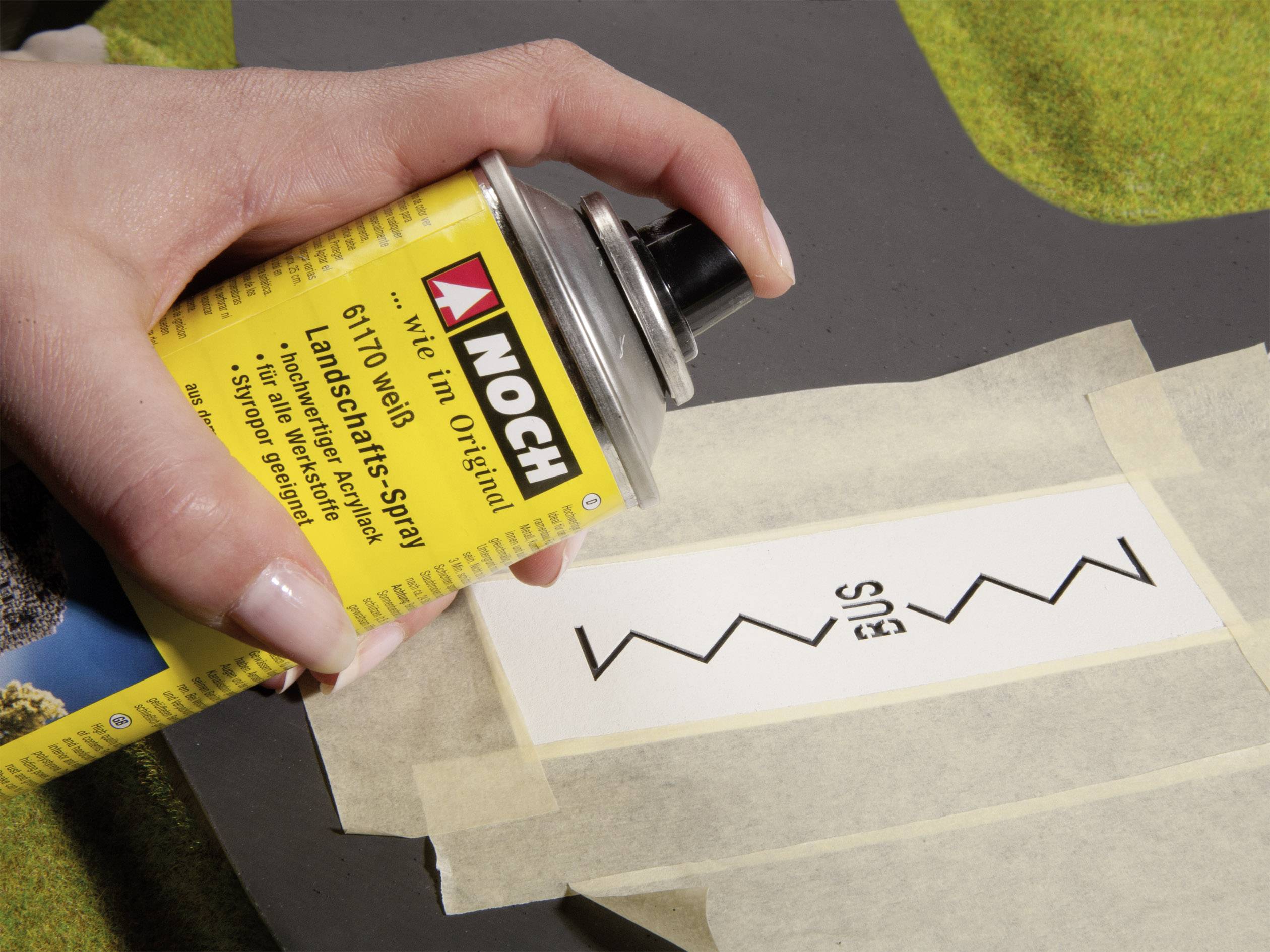 A hand holds a yellow spray can over a pattern masked with painter's tape on a surface, ready to spray.
