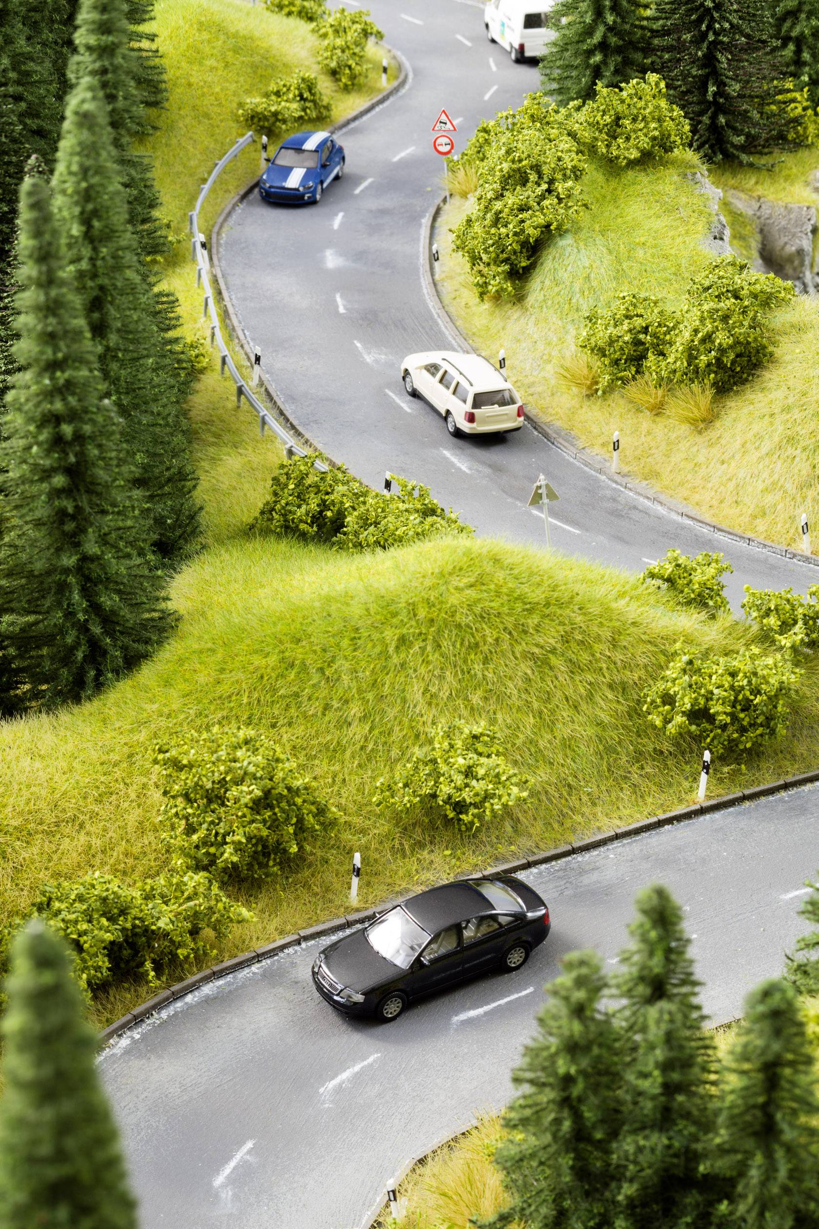 A winding country road in hilly, green countryside. Three cars are driving in the same direction. Road signs indicate upcoming bends.