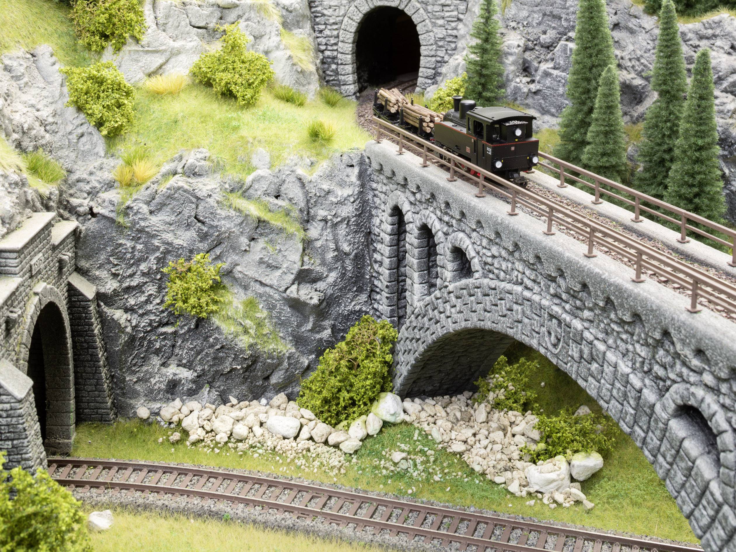A small model train travels across a stone bridge through a mountainous landscape with pine trees and a tunnel.