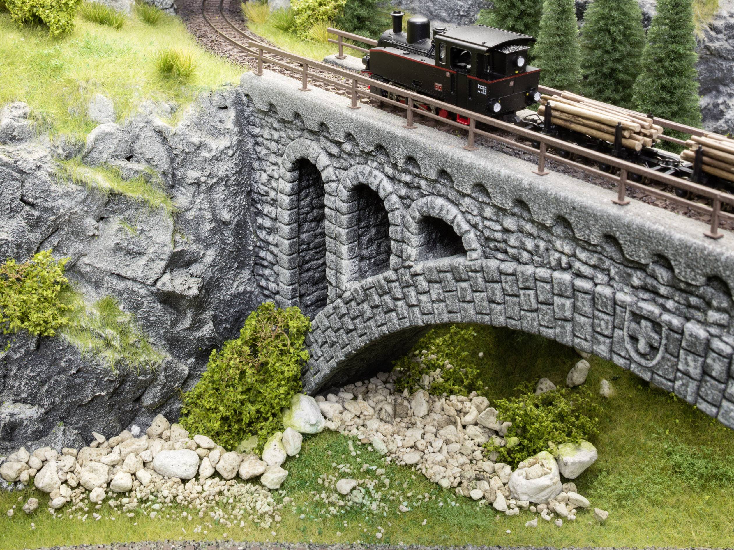 A small toy train travels across a stone bridge over a rocky stream in a miniature landscape with trees and grass.
