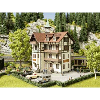 A model of a half-timbered house with a garden, surrounded by trees. People are standing in front of the building, and cars can be seen in the car park.