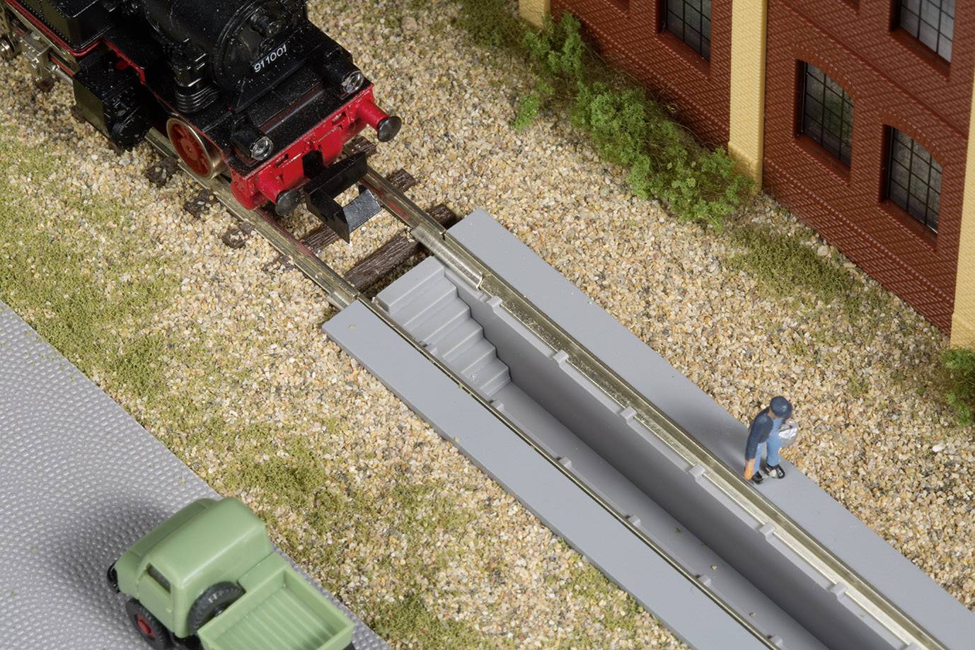 A model train with a red locomotive is travelling on tracks beside a building. A man is walking next to the tracks. A green car is standing in the foreground.