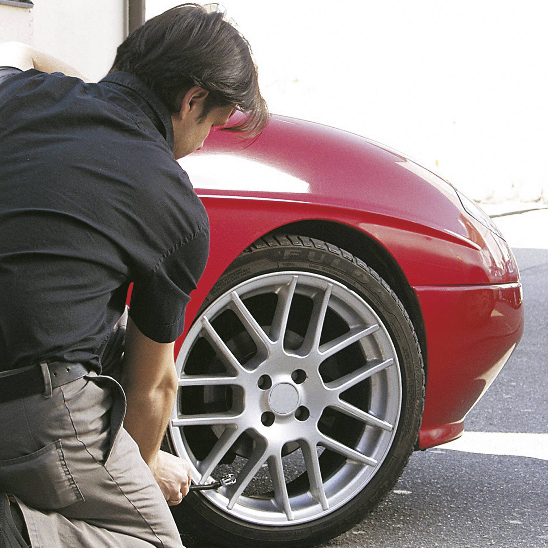A man is checking the tyre pressure of a red car in a car park.