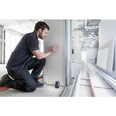 A man is kneeling and marking a line on a wall using a laser measuring device in a room under construction.
