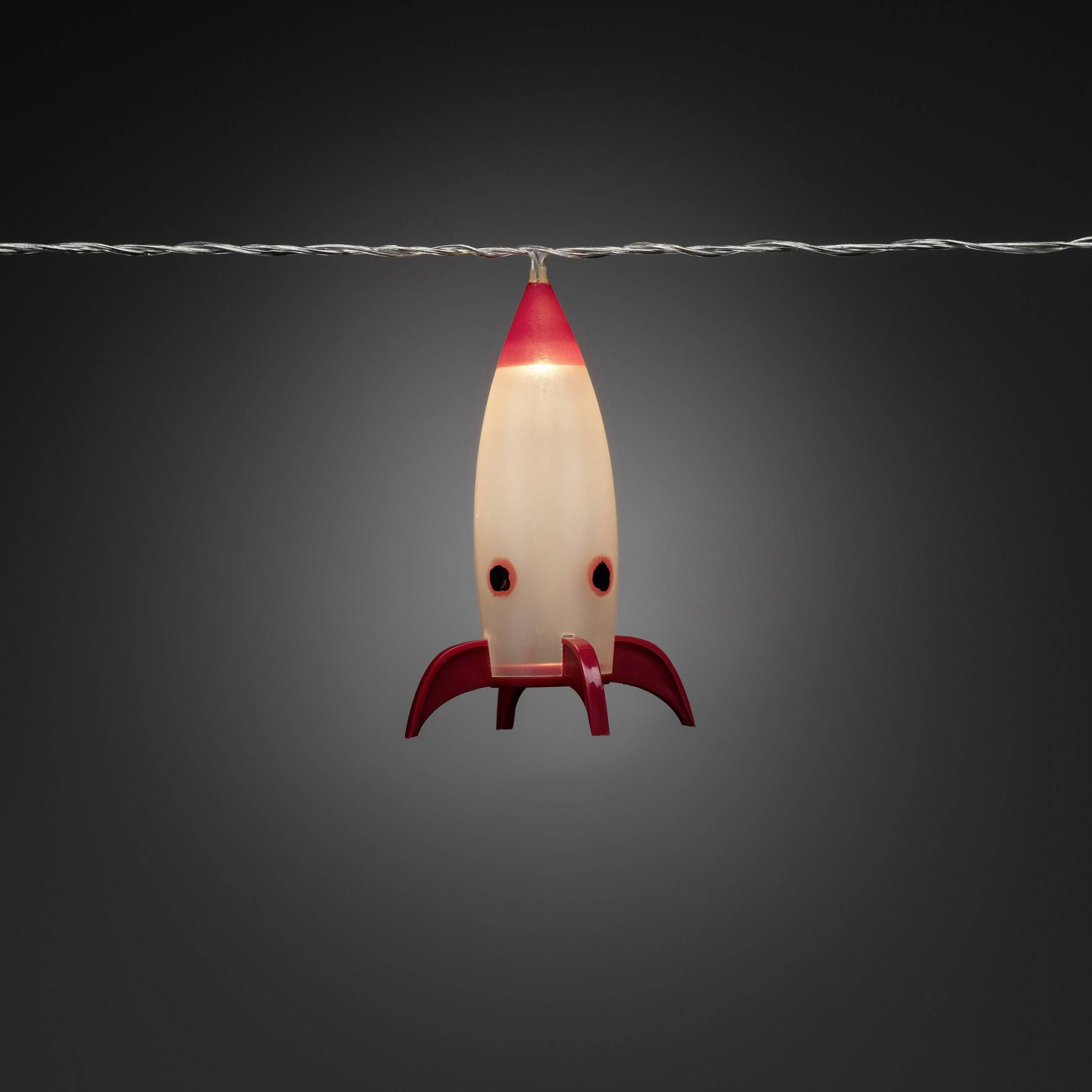 A rocket-shaped lamp with a luminous body and red details, hanging from a cable, against a grey background.