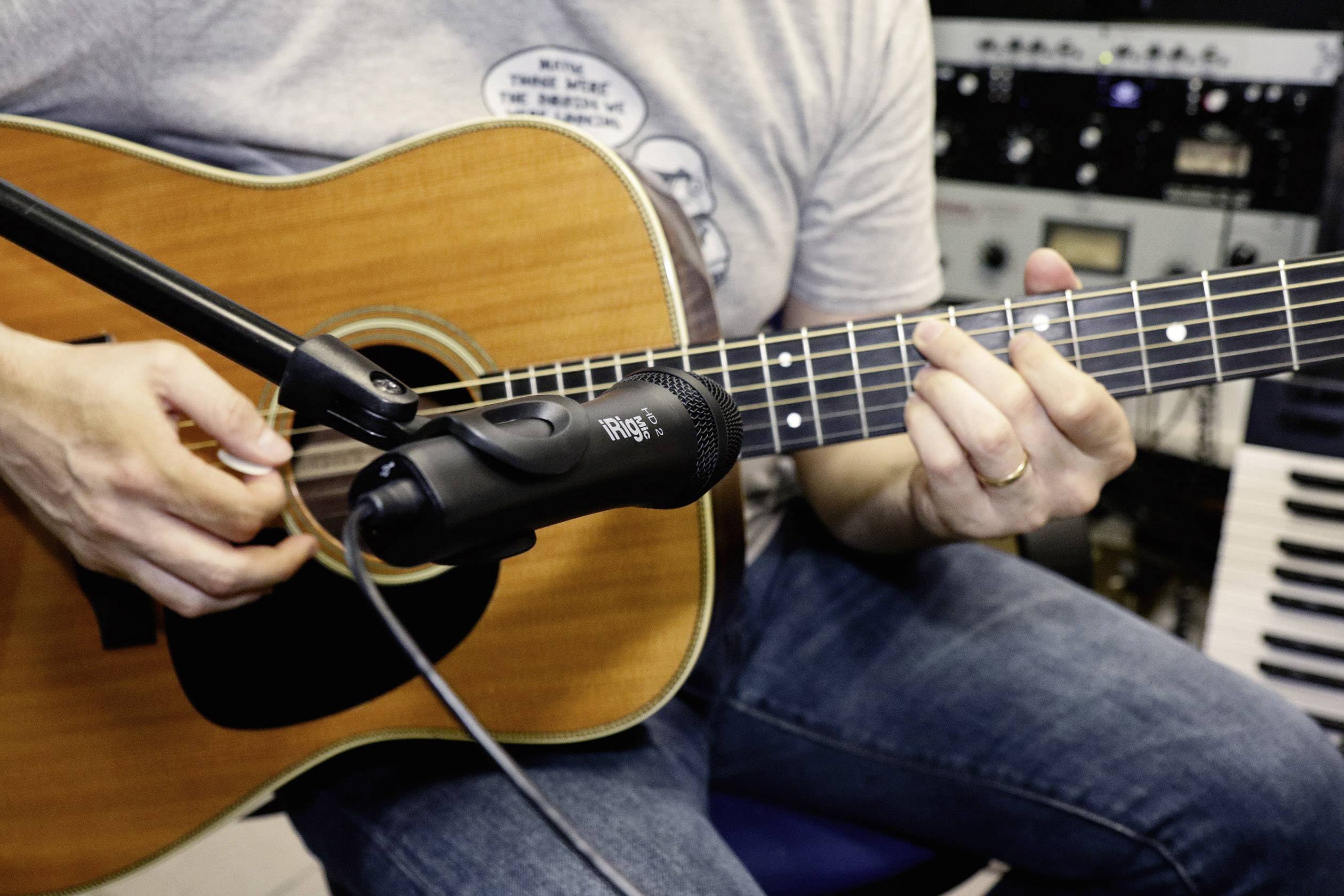 A person is playing an acoustic guitar while a microphone captures the sound. Studio equipment can be seen in the background.
