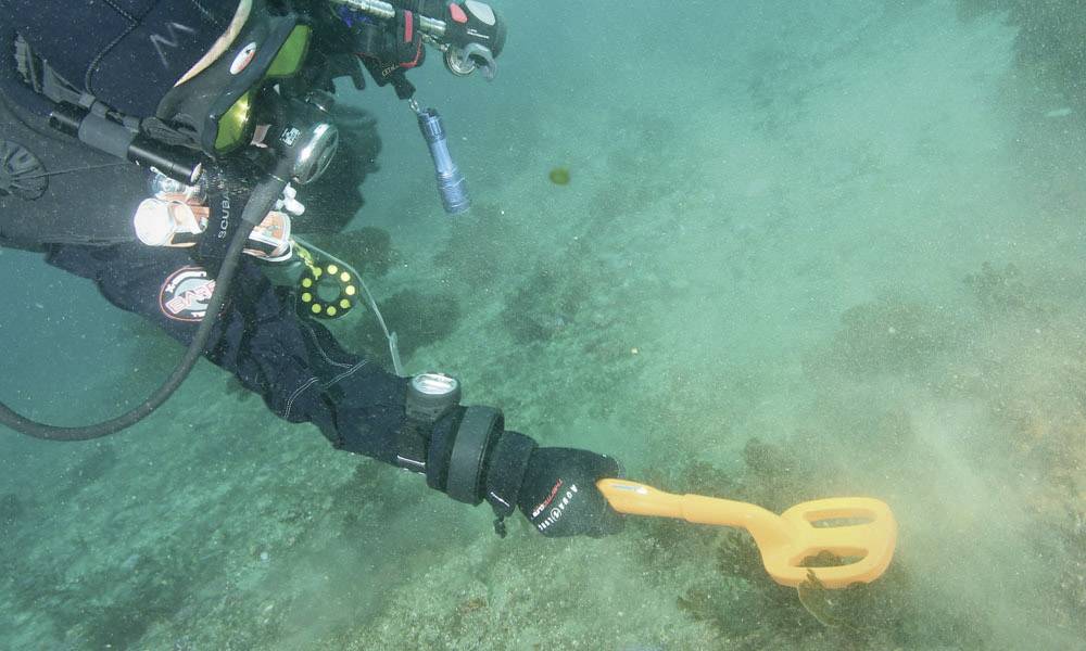 A diver is using a metal detector underwater to search for objects in the sand, surrounded by a blue underwater environment.