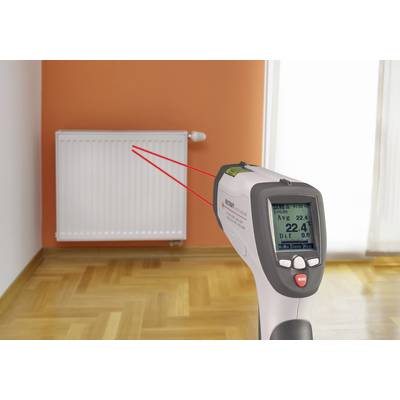 An infrared thermometer measures the surface temperature of a radiator in a room with a wooden floor and orange wall.