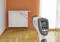 An infrared thermometer measures the surface temperature of a radiator in a room with a wooden floor and orange wall.